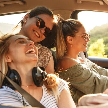 Infotage bei DEGENER Drei lachende Frauen sitzen dicht beieinander im Auto und genießen eine Roadtrip-Fahrt im warmen Sonnenlicht